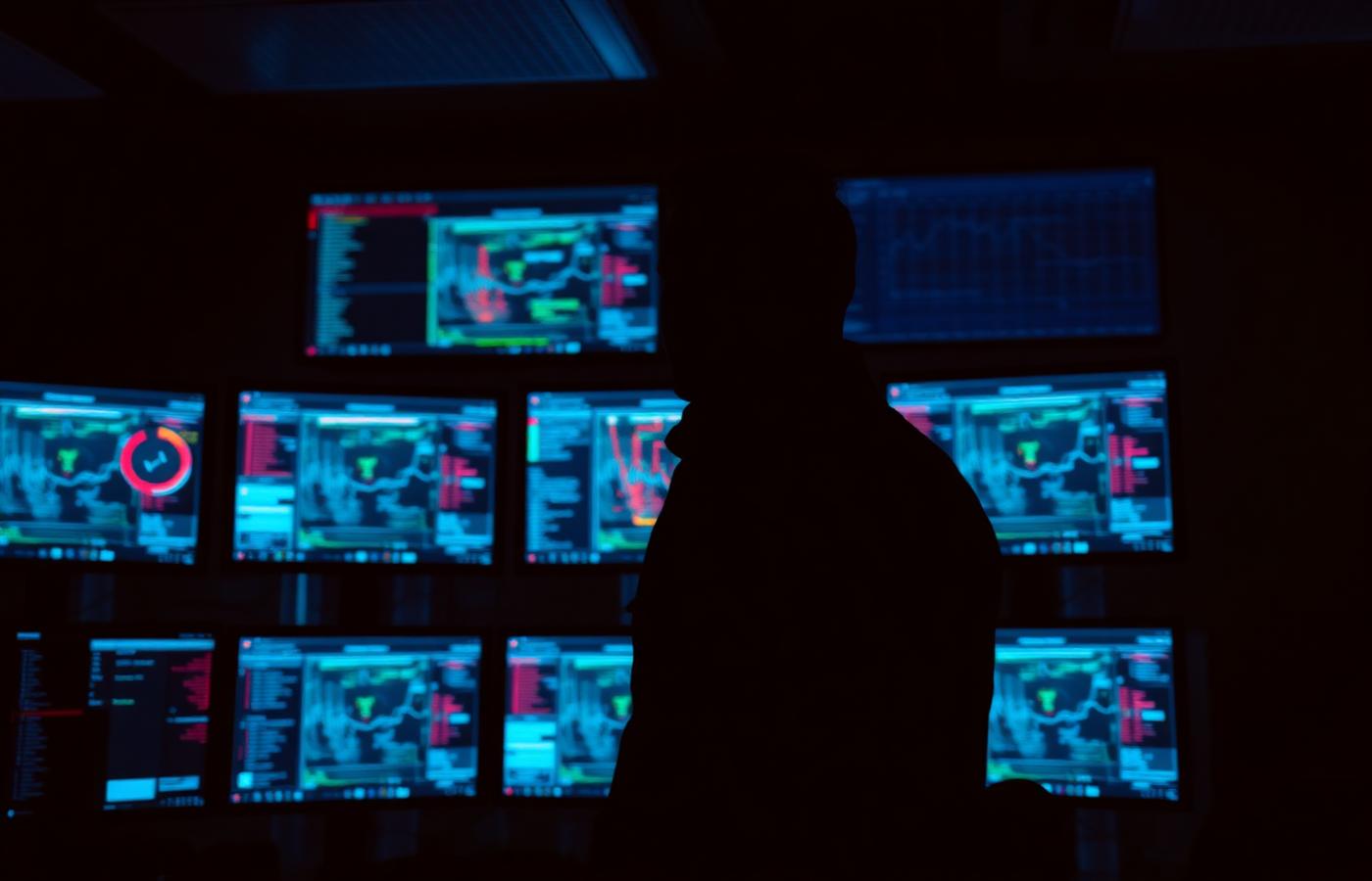 Silhouette of a security analyst in front of a wall of monitoring dashboards at night